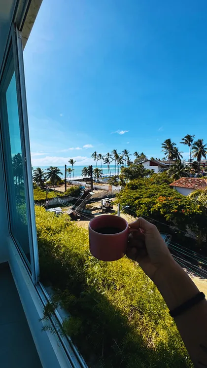 Mão segurando uma xícara de café em uma varanda com vista para coqueiros e o mar.