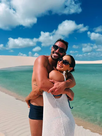 Juliana abraçada ao companheiro em uma praia de águas claras e dunas.